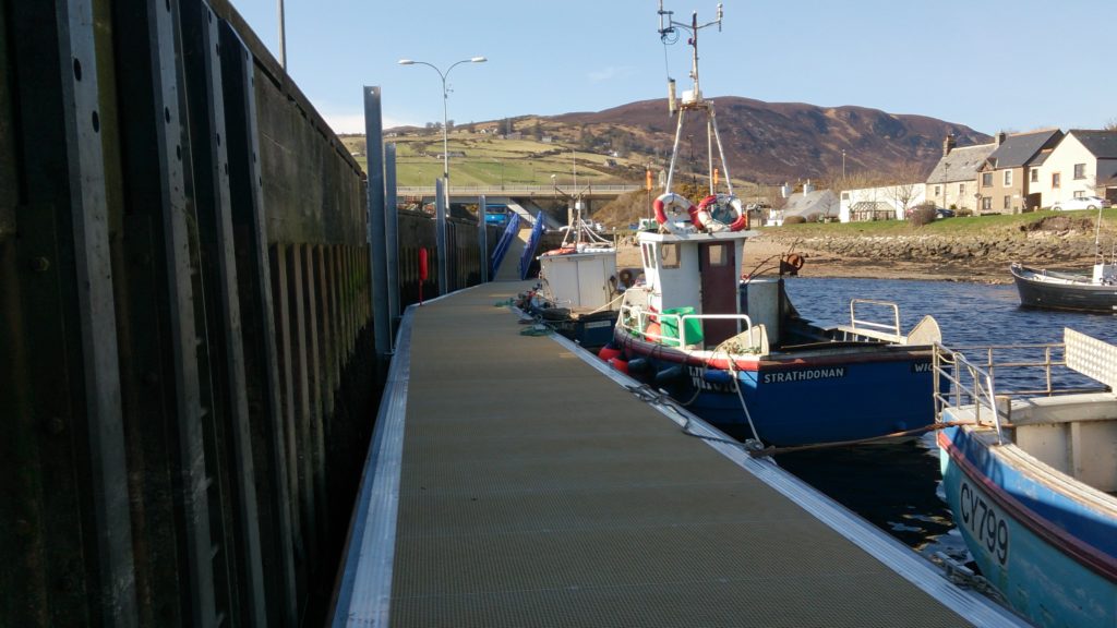 Pontoon curving alongside a quay wall with colourful fishing vessels moored
